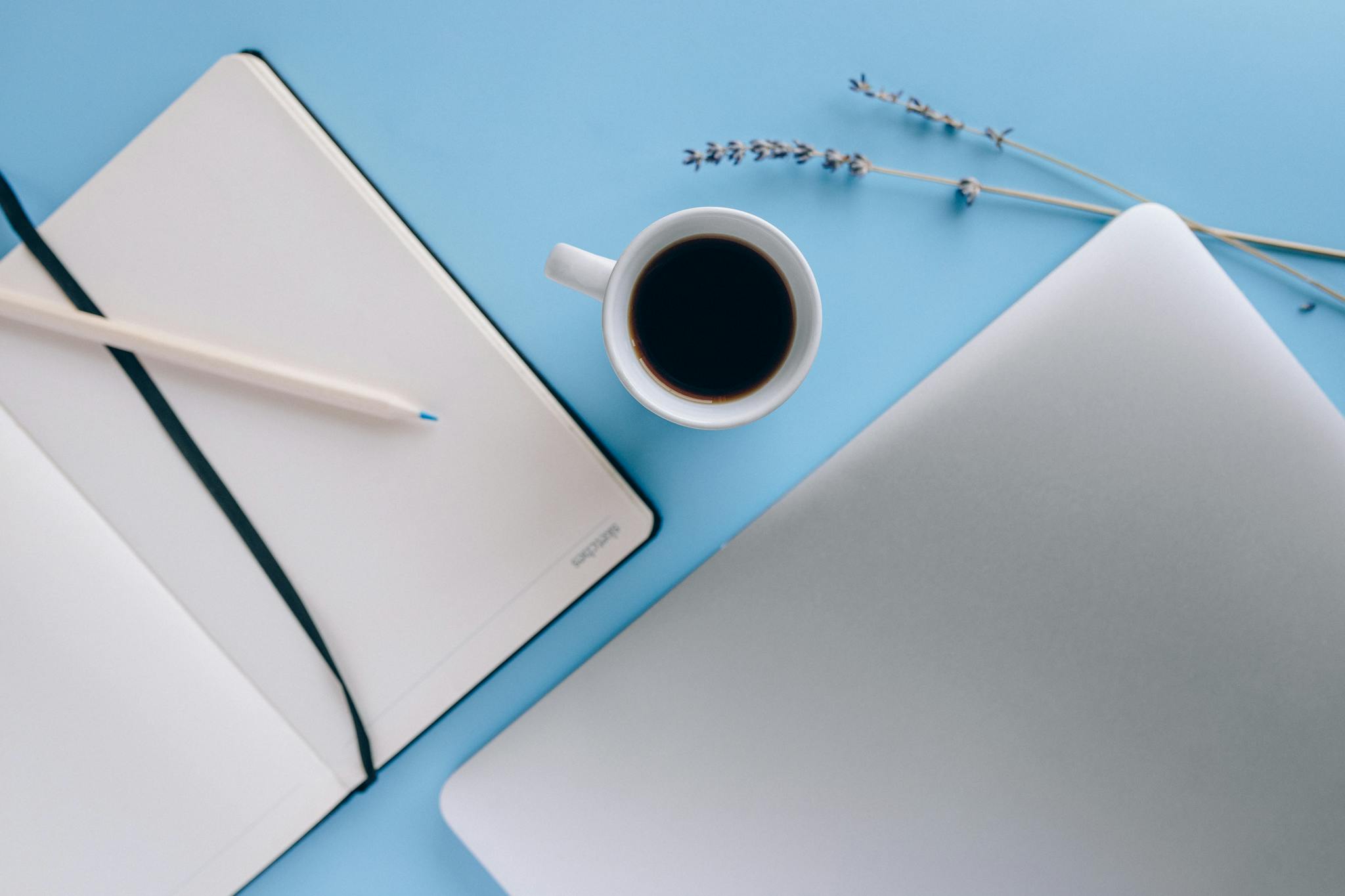 Top view of a minimal workspace featuring a notebook, coffee, and lavender on a blue background.